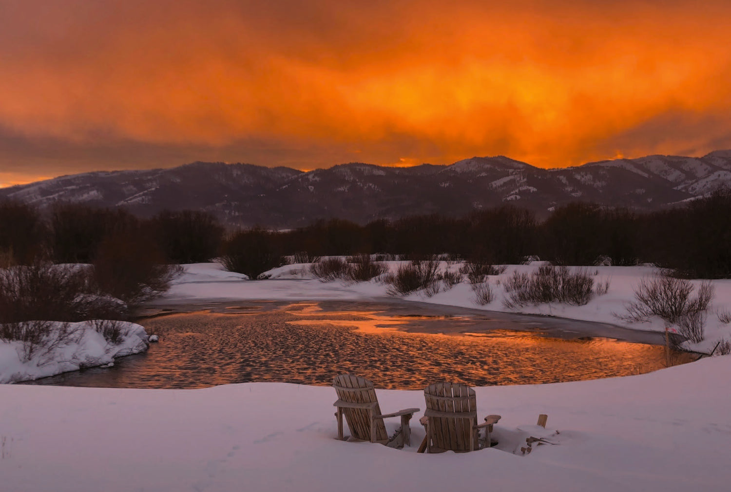 Two Adirondack chairs in the snow beside the frozen pond under a burning orange sunset